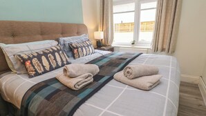 A neatly made bed with a checkered bedspread and two rolled towels, a nightstand with a lamp, and a window with curtains.