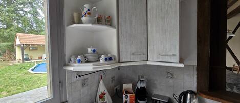 A kitchen with white cabinetry, a built-in shelf with white and blue floral dishware, a coffee maker, a kettle, and a small window overlooking a backyard.