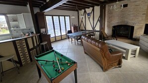 Een ruime woonkamer met een pooltafel, een bank, een eettafel met stoelen, een open haard en een keukenhoek.