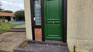 Een groene deur met een bakstenen basis, een klein raam met bloemenmotief, en een betonnen pad dat naar een gebouw met pannendak leidt.