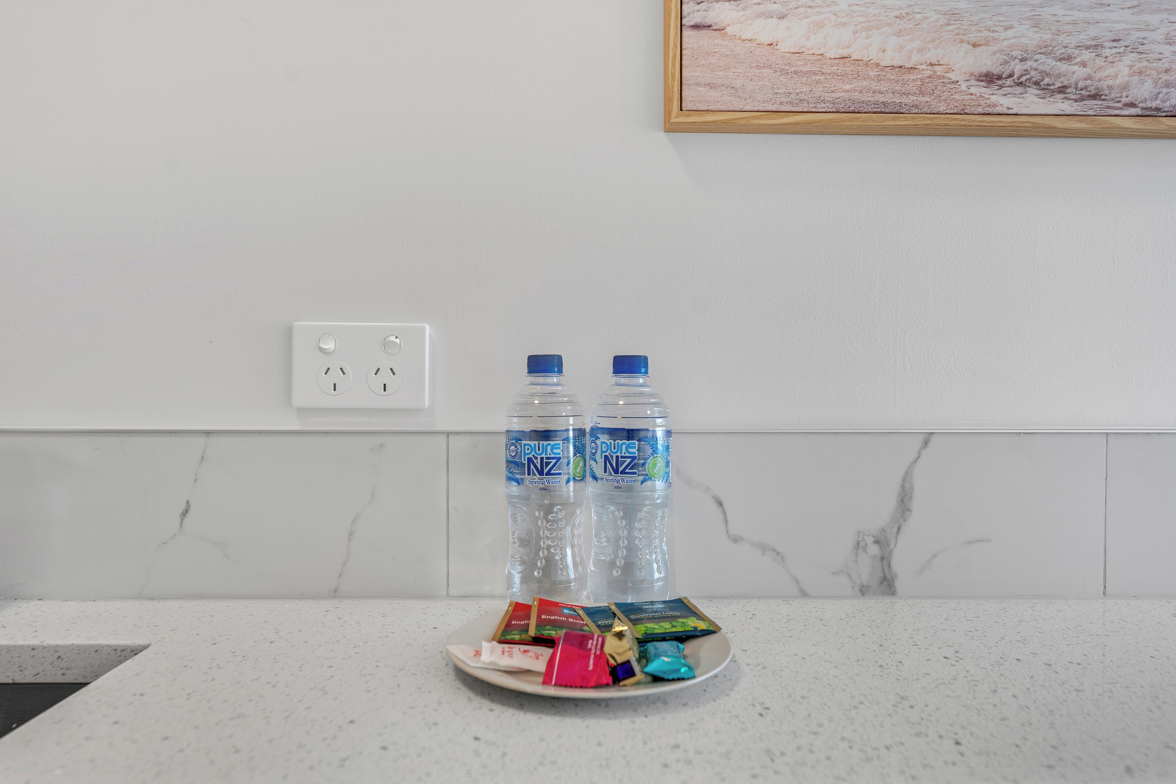 Un comptoir de cuisine avec deux bouteilles d’eau, une assiette contenant des portions de nourriture emballées et une prise électrique sur un mur recouvert de carreaux blancs.