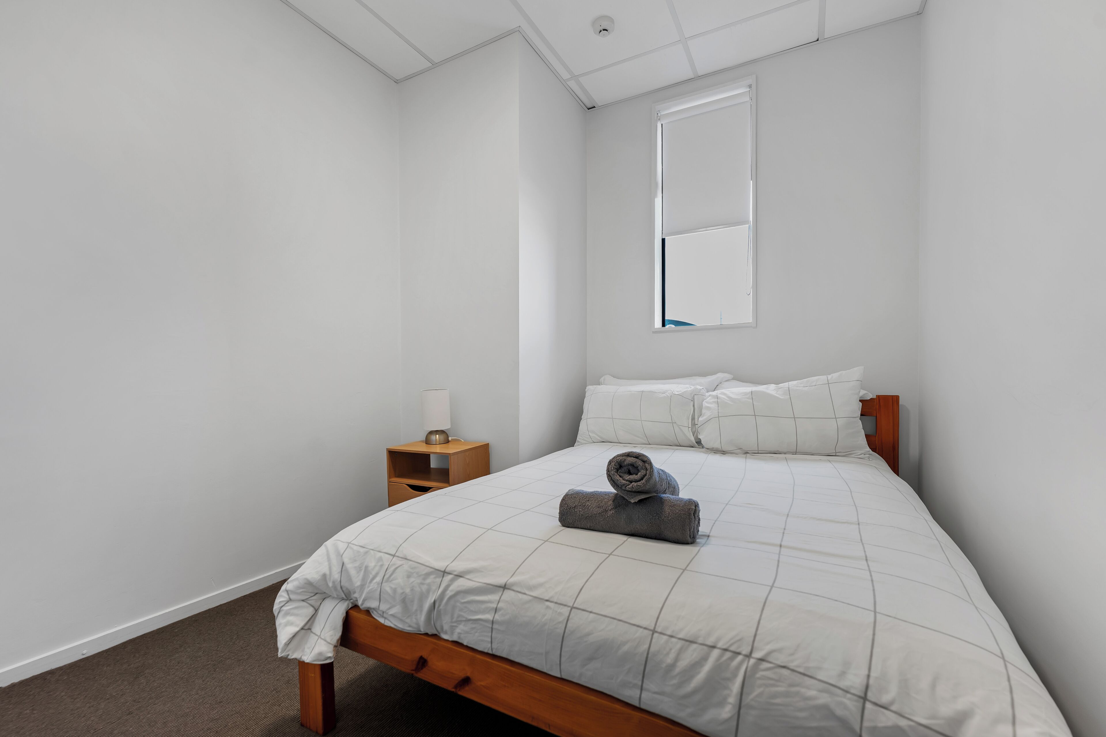 Een slaapkamer met een bed, nachtkastje, raam en een spiegel aan de muur.