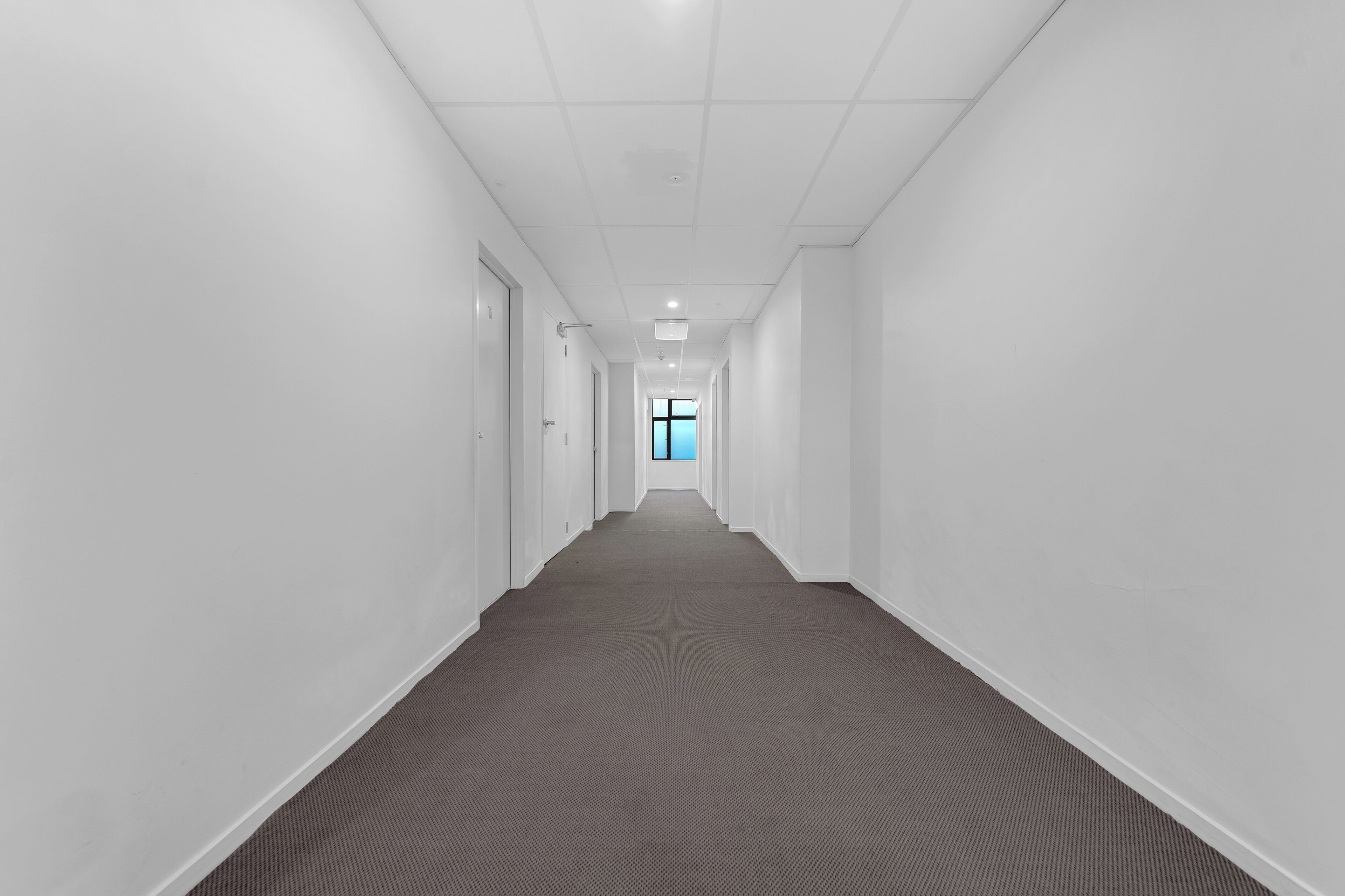 A long, narrow hallway with white walls and a carpeted floor, leading to a door at the end.