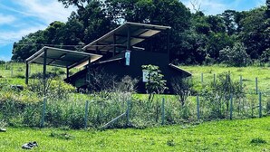 A small, open-air structure with a metal roof and walls, surrounded by a grassy field and a fence. - Cozy farm in front of the dam, for water sports and fishing. (Serrinha Bragança Paulista)