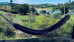 A hammock in a lush outdoor setting with a view of a lake and distant hills. - Cozy farm in front of the dam, for water sports and fishing. (Serrinha Bragança Paulista)