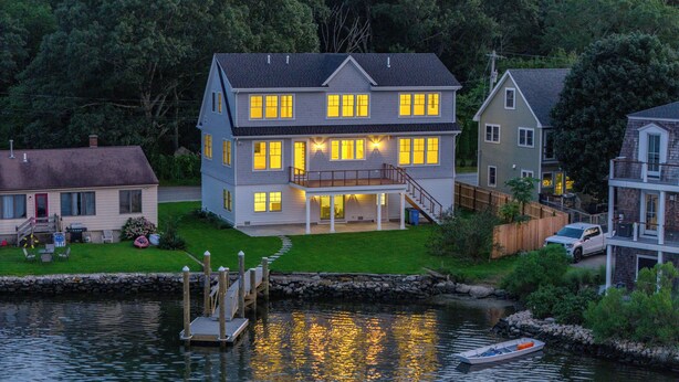 A large two-story house with a dock extending into a body of water, surrounded by greenery and other houses. - Luxury Waterfront Home Private Dock, Stunning Views & Sleeps 14 (South Kingstown)