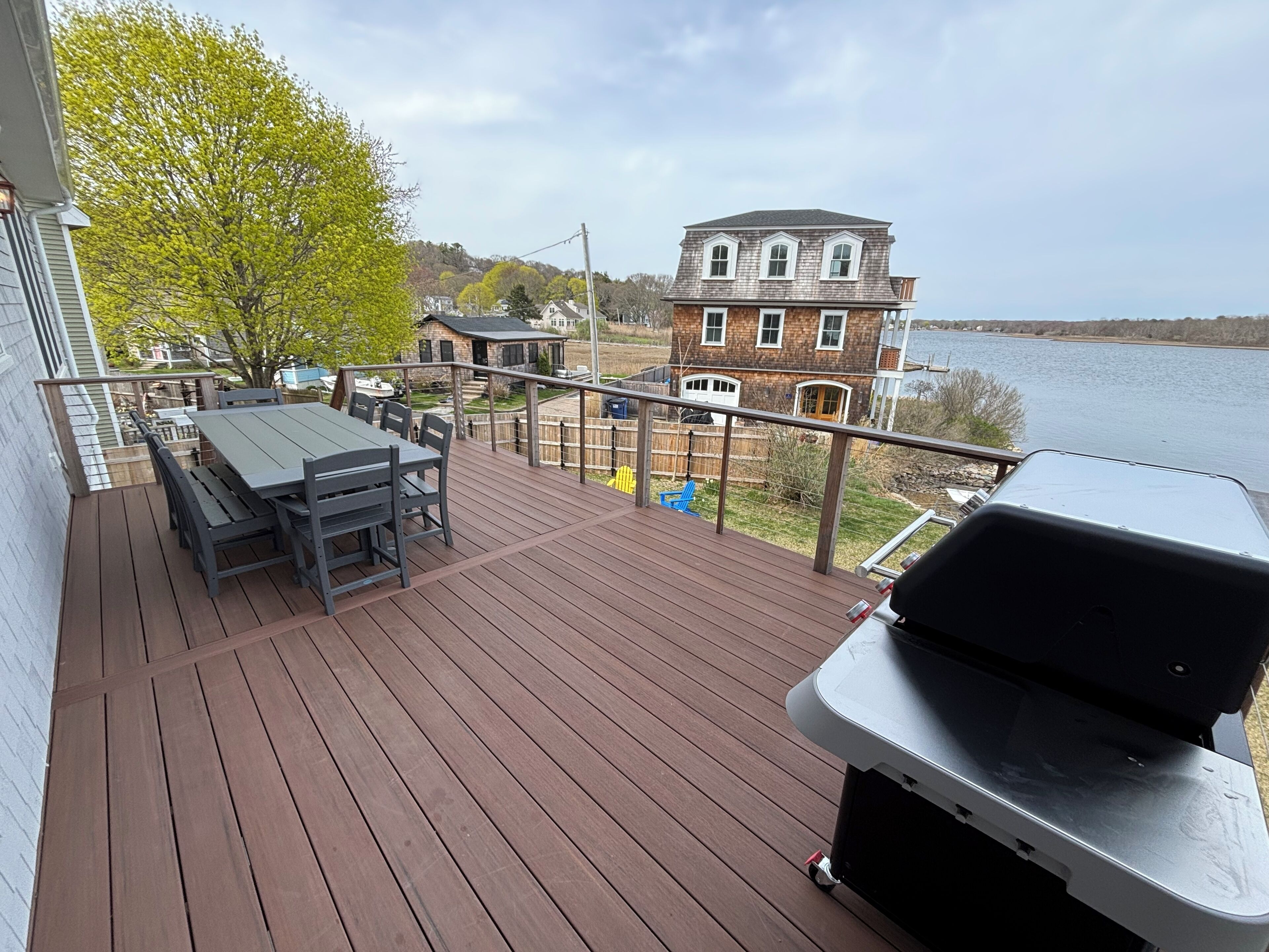 A wooden deck with a grill and table, overlooking a lake and houses.
