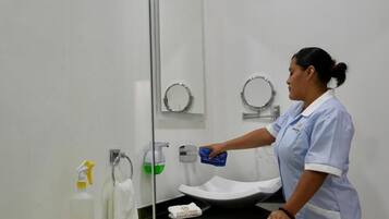 Un baño con inodoro, lavamanos y espejo. Una persona está limpiando la zona del lavamanos.