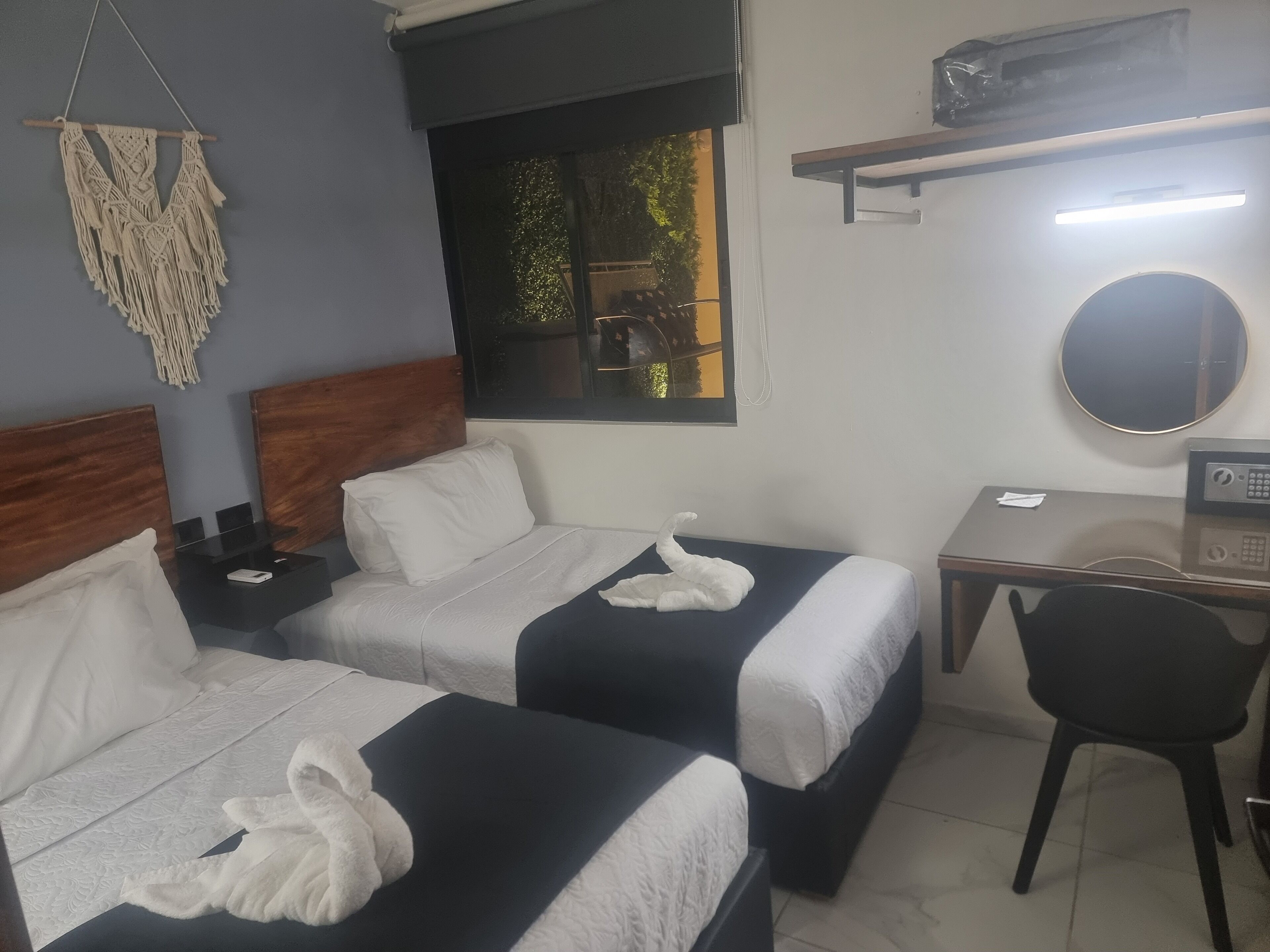 A hotel room with two beds, a desk, a chair, a macrame wall hanging, and a window with a view of greenery.