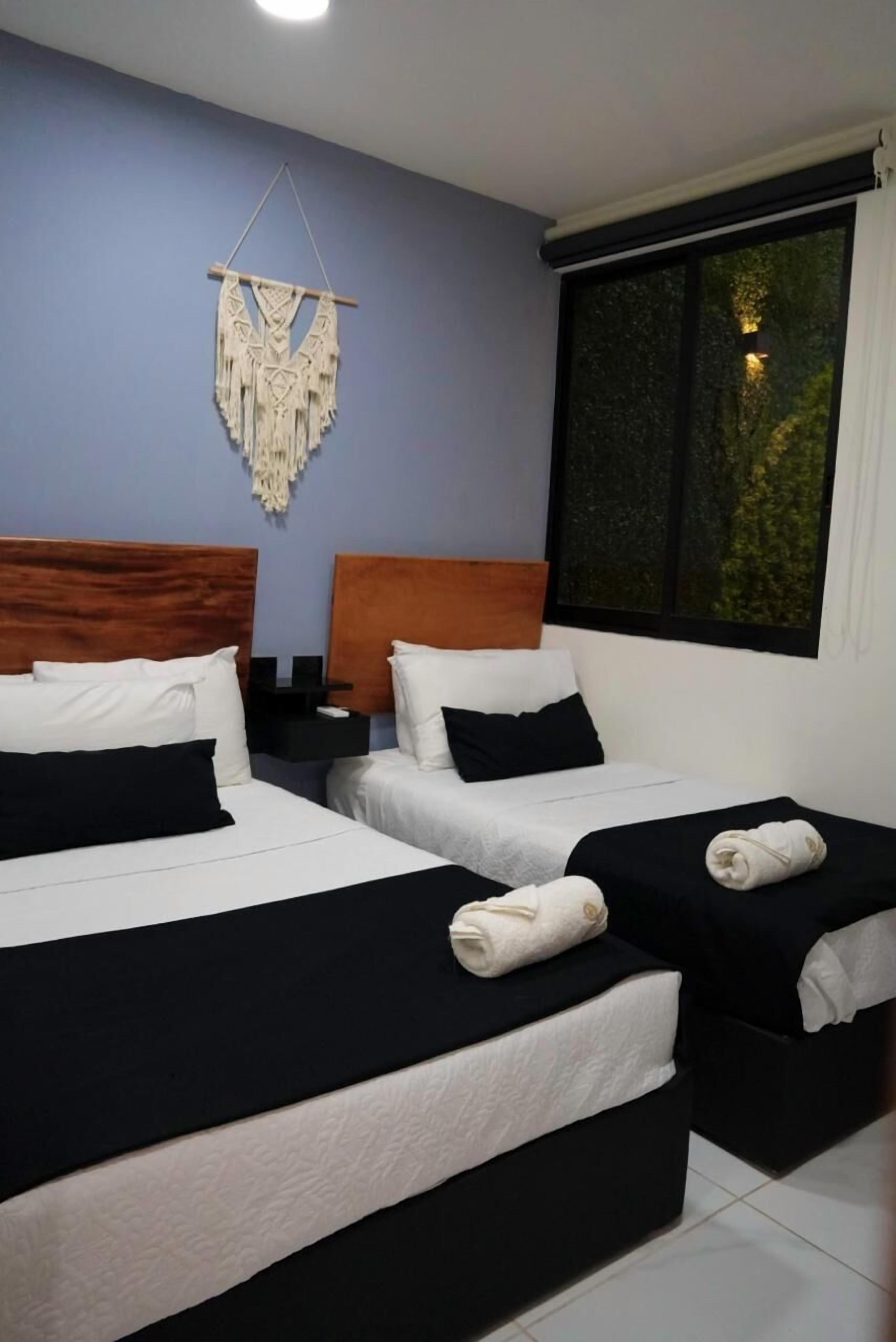 Habitación de hotel con dos camas, una mesita de noche, un tapiz de macramé y una ventana con vista a árboles.