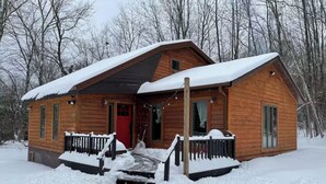 A wooden cabin with a red door, a porch, and a small deck, surrounded by snow and trees.