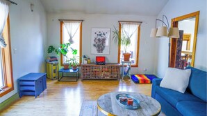 A spacious living room with a blue sofa, a coffee table, and a wooden floor. There are two windows with curtains, a bookshelf, and a potted plant.