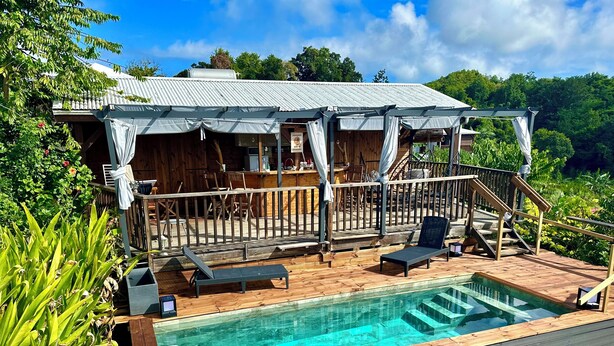 A wooden deck with a swimming pool, lounge chairs, and a covered seating area with a view of the surrounding greenery. - Gîte Santa Maria (Grand-Bourg)