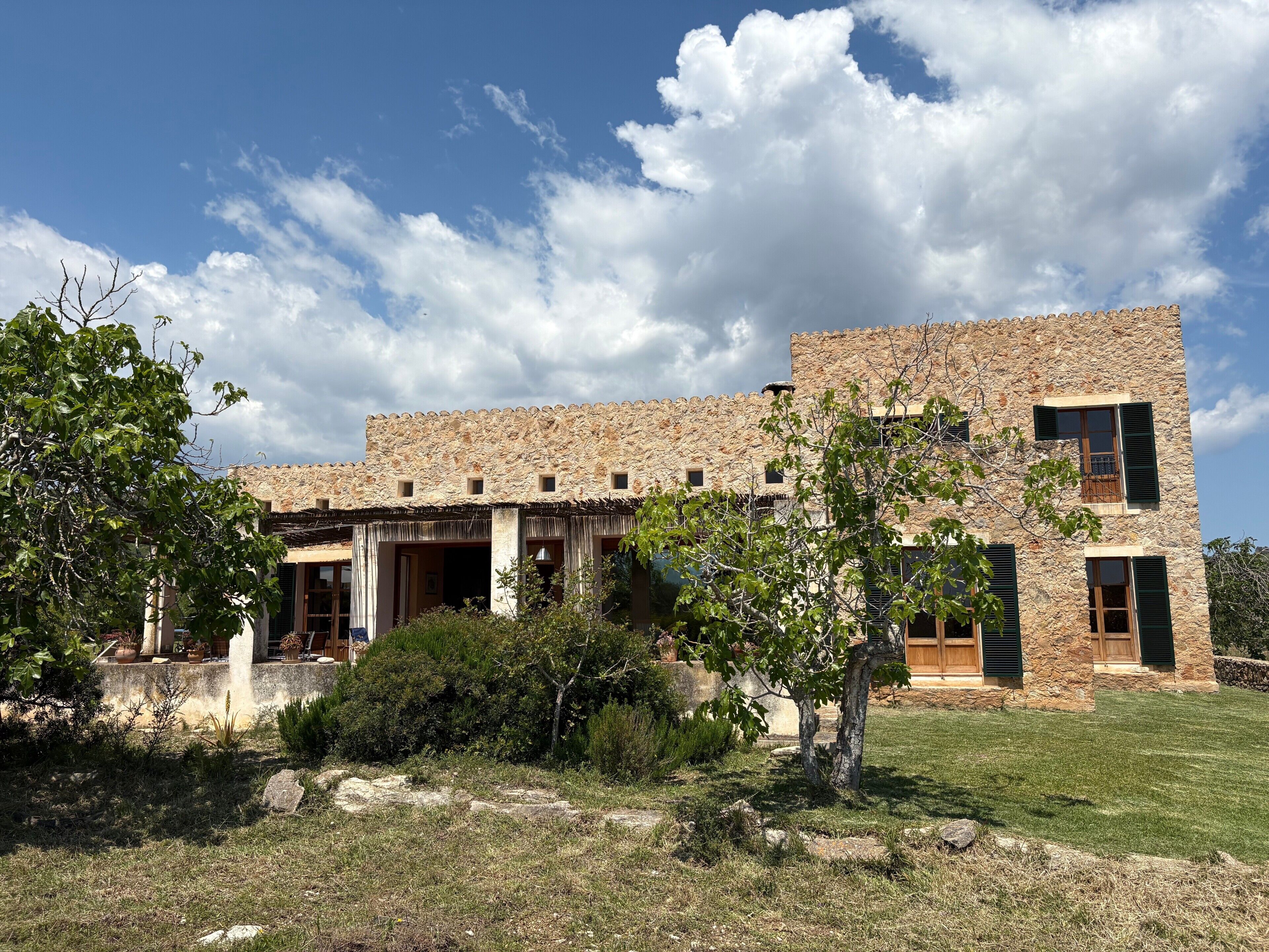 A stone building with multiple windows and a door, surrounded by greenery and a clear sky.