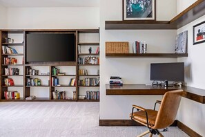 Ein Wohnzimmer mit einem großen Flachbildfernseher, einer Bücherwand, einem Schreibtisch mit Computer und einem Ledersessel.