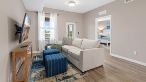 A living room with a sofa, ottoman, TV, and a window with curtains. - Gulf Access with Boat Slip, Heated Saltwater Pool, Dog-Friendly - Bacino Vista #204 - Roelens (Cape Coral)