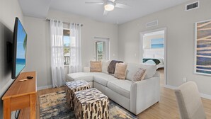 A modern living room with a sectional sofa, a flat-screen TV, a ceiling fan, and a view of the ocean through a door. - Gulf Access with Boat Slip, Heated Saltwater Pool, Dog-Friendly - Bacino Vista #207 - Roelens (Cape Coral)