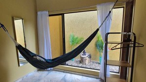 Une pièce avec un hamac, une fenêtre avec des rideaux, une petite plante et une étagère en métal.