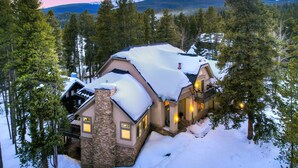 A snow-covered lodge with a stone chimney, a balcony, and a view of snow-capped mountains. - Lakeshore Lodge | Private Hot Tub, Movie Room, & Views for Family Gatherings! (Blue River)