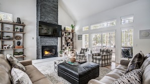 A spacious living room with a stone fireplace, comfortable seating, and large windows. - Lakeshore Lodge | Private Hot Tub, Movie Room, & Views for Family Gatherings! (Blue River)