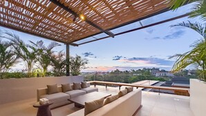 A covered outdoor seating area with a sofa and a table, surrounded by plants and overlooking a cityscape.