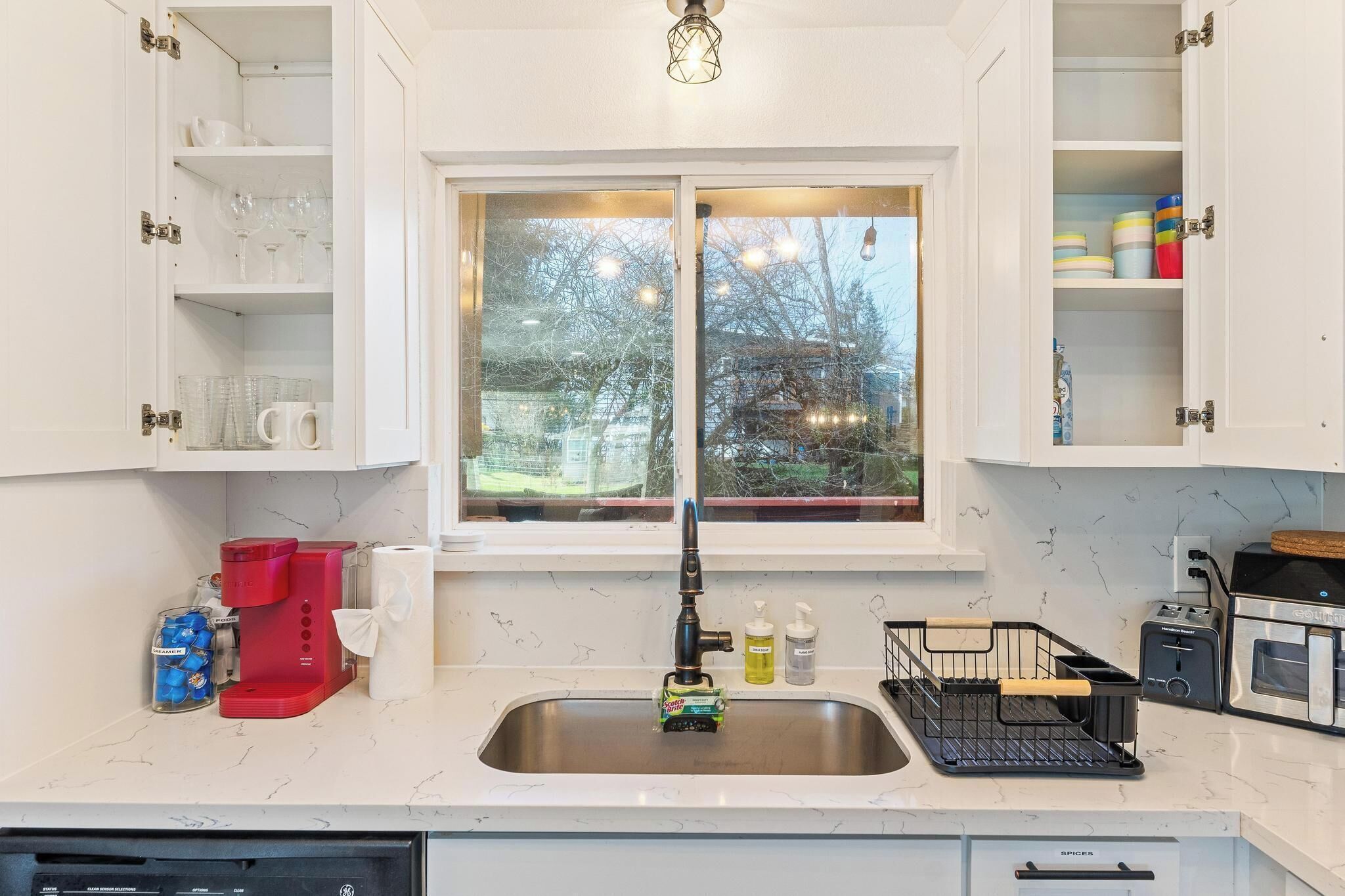 A kitchen with a double sink, a coffee maker, and a window with a view of trees and buildings.