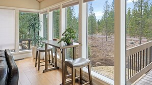 A sunlit room with a wooden table and two stools, large windows with a view of trees, and a black leather couch. - 20 Aspen - Elegant & Updated & Perfect for Family! SHARC, Hot Tub (Bend)