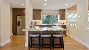 A modern kitchen with wooden cabinets, a stainless steel refrigerator, a large island with bar stools, and a window providing a view of the outdoors. - 20 Aspen - Elegant & Updated & Perfect for Family! SHARC, Hot Tub (Bend)