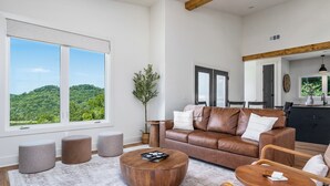 Un salón espacioso con un gran ventanal, un sofá de cuero marrón, una mesa de centro redonda y vistas a un paisaje verde.