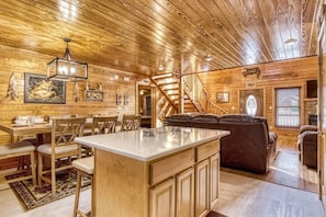 Interior de una cabaña de troncos con comedor, sala de estar con chimenea y una escalera.
