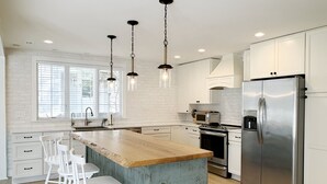 A modern kitchen with white cabinetry, a wooden island with bar stools, stainless steel appliances, and pendant lighting. - Cape Cod Charm -- Steps From Beach, 3 King Beds, Beach Equipment & Linens (Lewes)