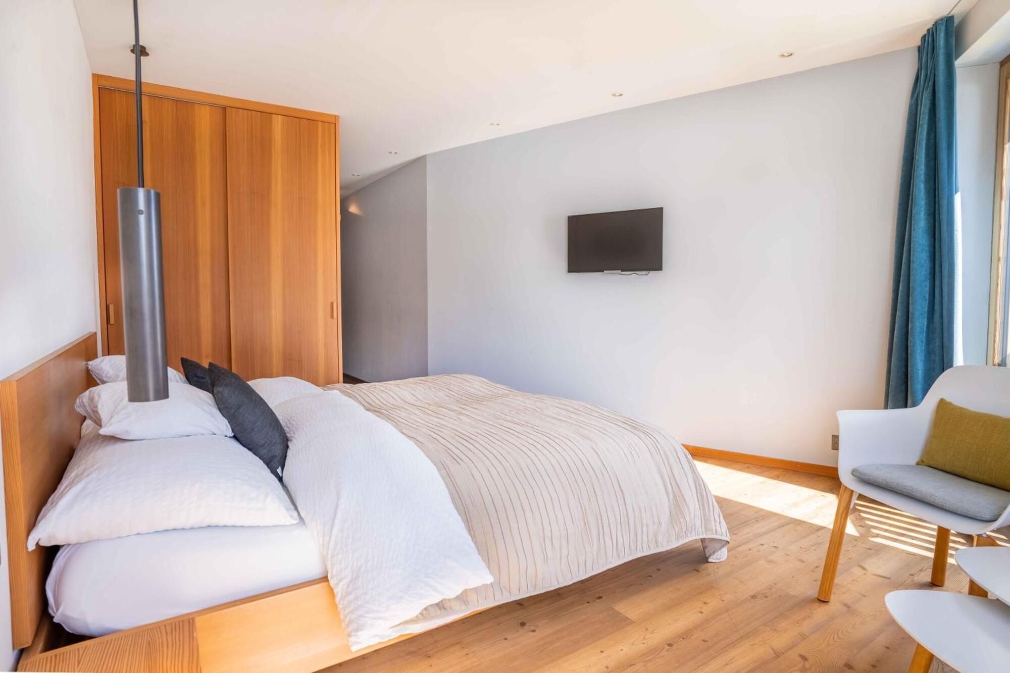 Un dormitorio moderno con cabecera de madera, cama con ropa blanca, televisor montado y un área de descanso con una silla y una mesa.