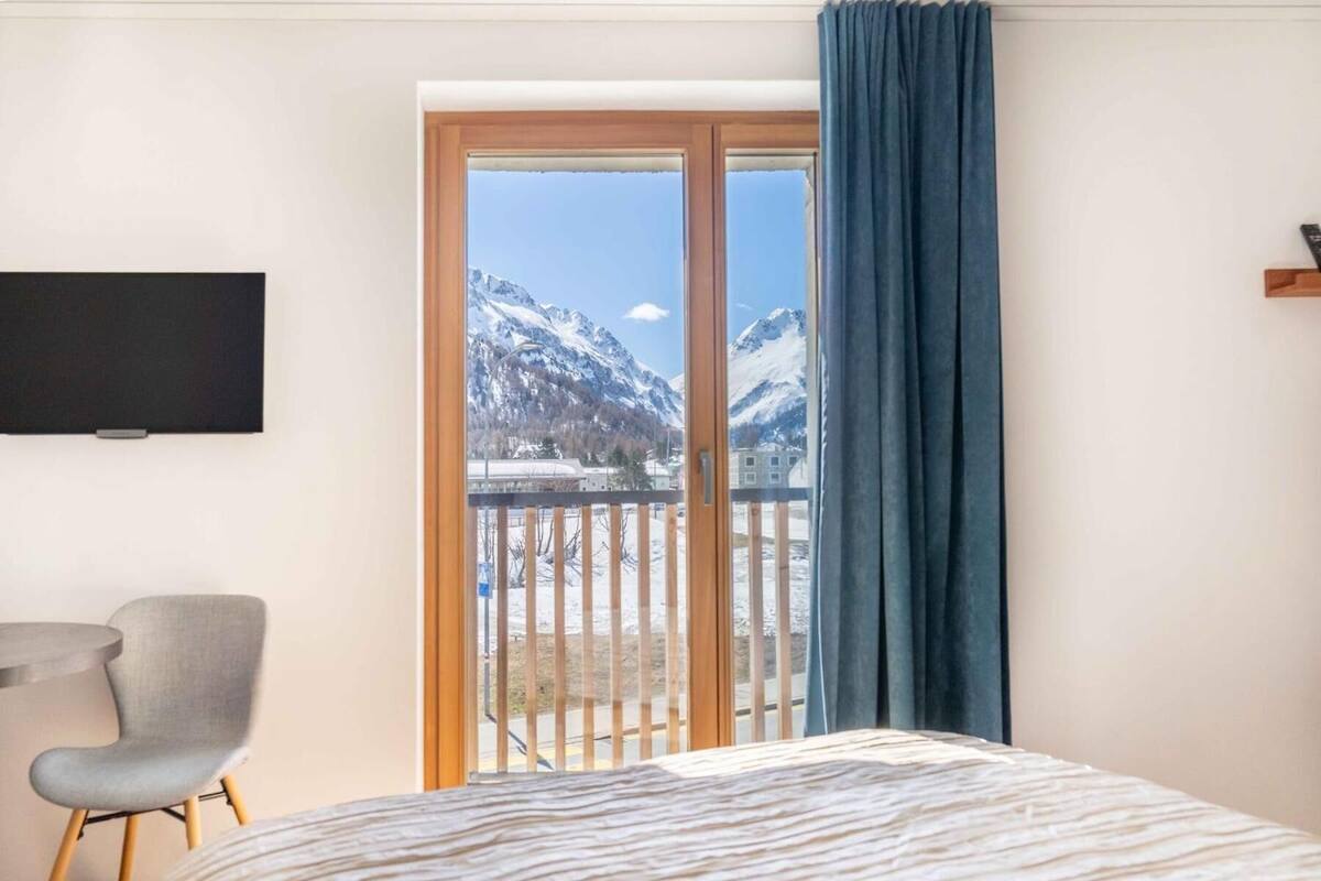 A hotel room with a bed, a chair, a TV, and a balcony with a mountain view.