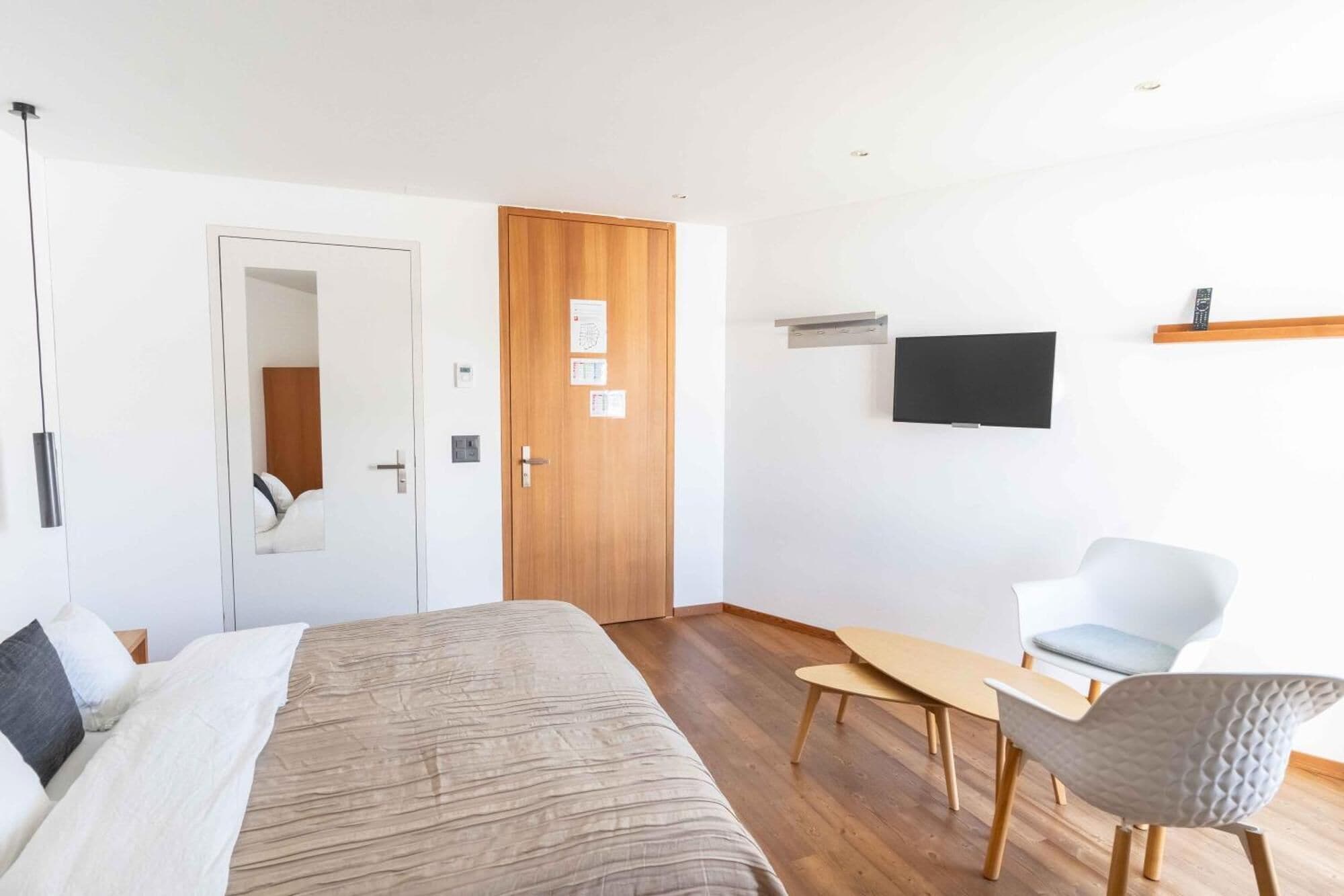 Moderni hotellihuone, jossa on sänky, puuovi, seinälle asennettu televisio sekä oleskelualue, jossa on pöytä ja tuolit.