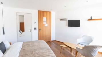 Moderni hotellihuone, jossa on sänky, puuovi, seinälle asennettu televisio sekä oleskelualue, jossa on pöytä ja tuolit.