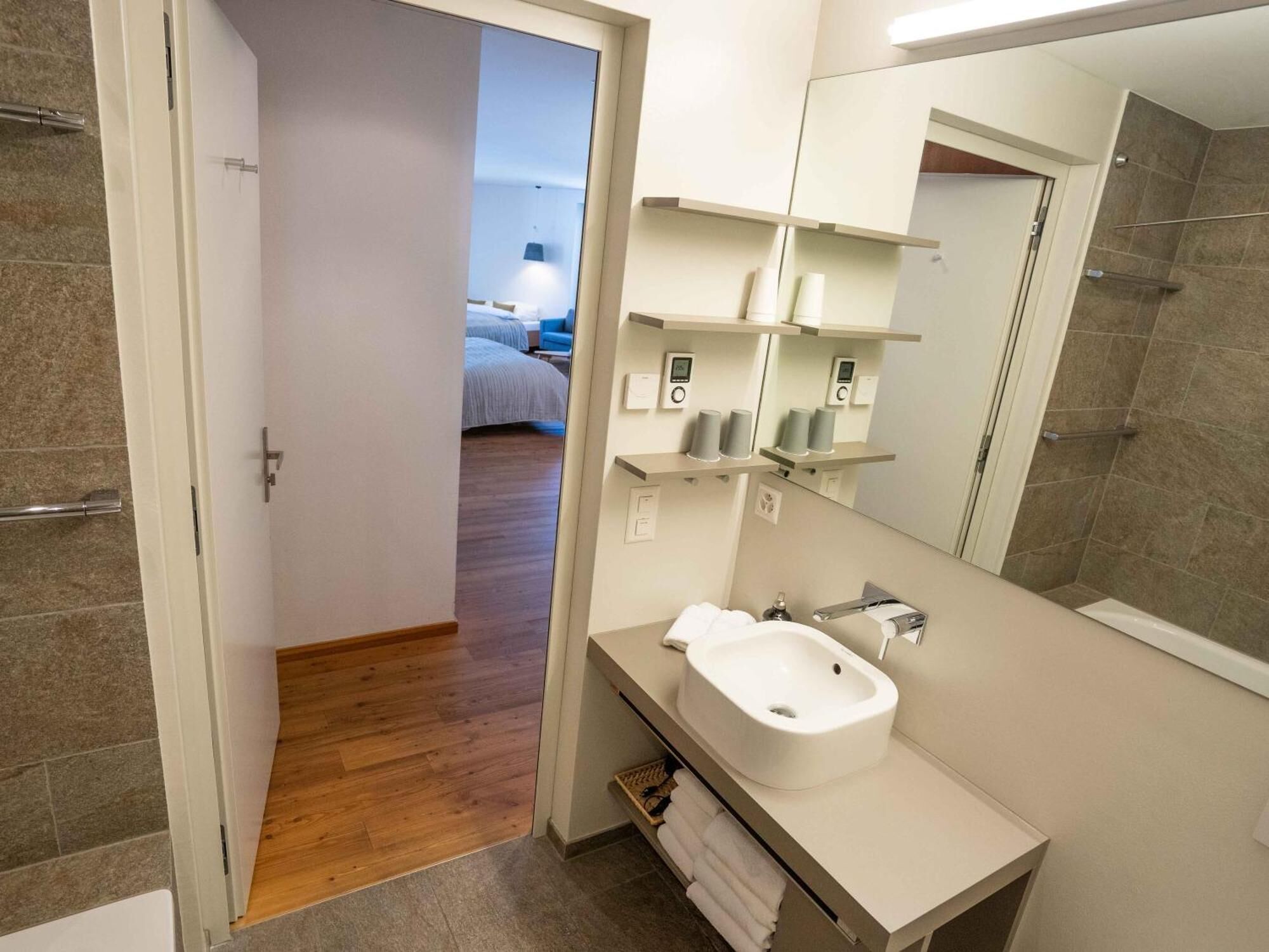 Un baño moderno con un lavabo blanco, espejo y estantes con artículos de aseo. Se observa un suelo de madera y una puerta que conduce a un dormitorio.