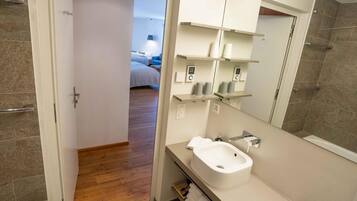 Un baño moderno con un lavabo blanco, espejo y estantes con artículos de aseo. Se observa un suelo de madera y una puerta que conduce a un dormitorio.