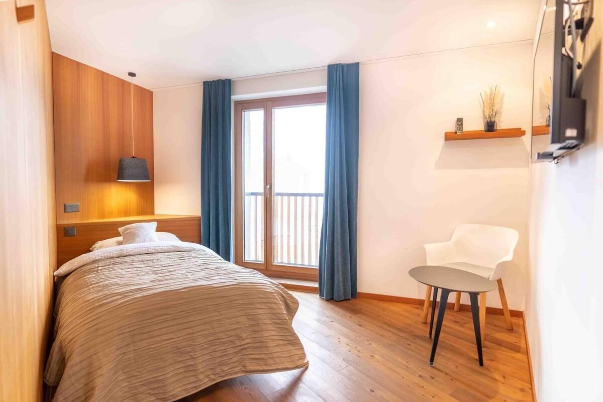 A hotel room with a bed, a chair, a small table, a window with curtains, and a wall-mounted shelf with a vase.