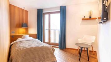A hotel room with a bed, a chair, a small table, a window with curtains, and a wall-mounted shelf with a vase.