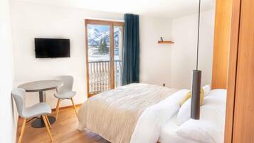 Une chambre d’hôtel comprenant un lit, un bureau avec une chaise, une télévision et un balcon offrant une vue sur des montagnes enneigées.