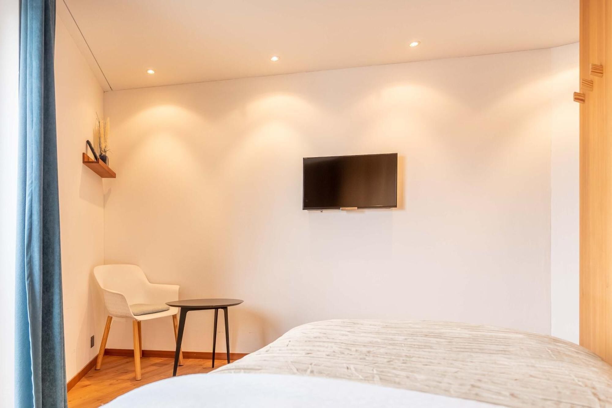 Ein schlicht eingerichtetes Hotelzimmer mit einem Bett, einem kleinen Tisch, einem Sessel und einem an der Wand montierten Fernseher.