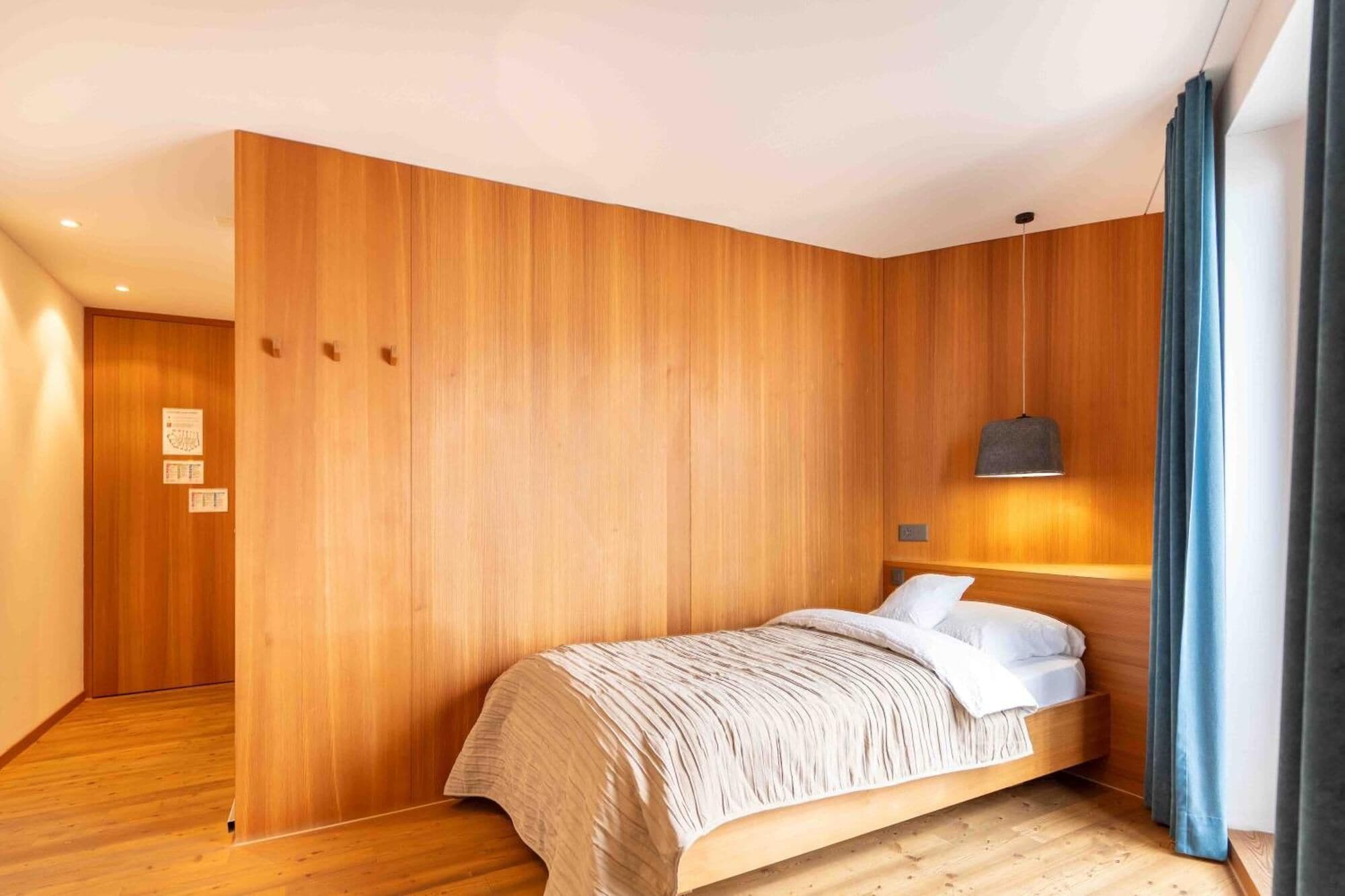 Habitación de hotel moderna con paredes de paneles de madera, una cama con colcha a rayas, una lámpara colgante y vistas al pasillo.
