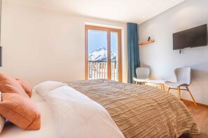 A hotel room with a bed, a chair, a TV, and a view of snow-capped mountains through a large window. - Longhin (Bregaglia)