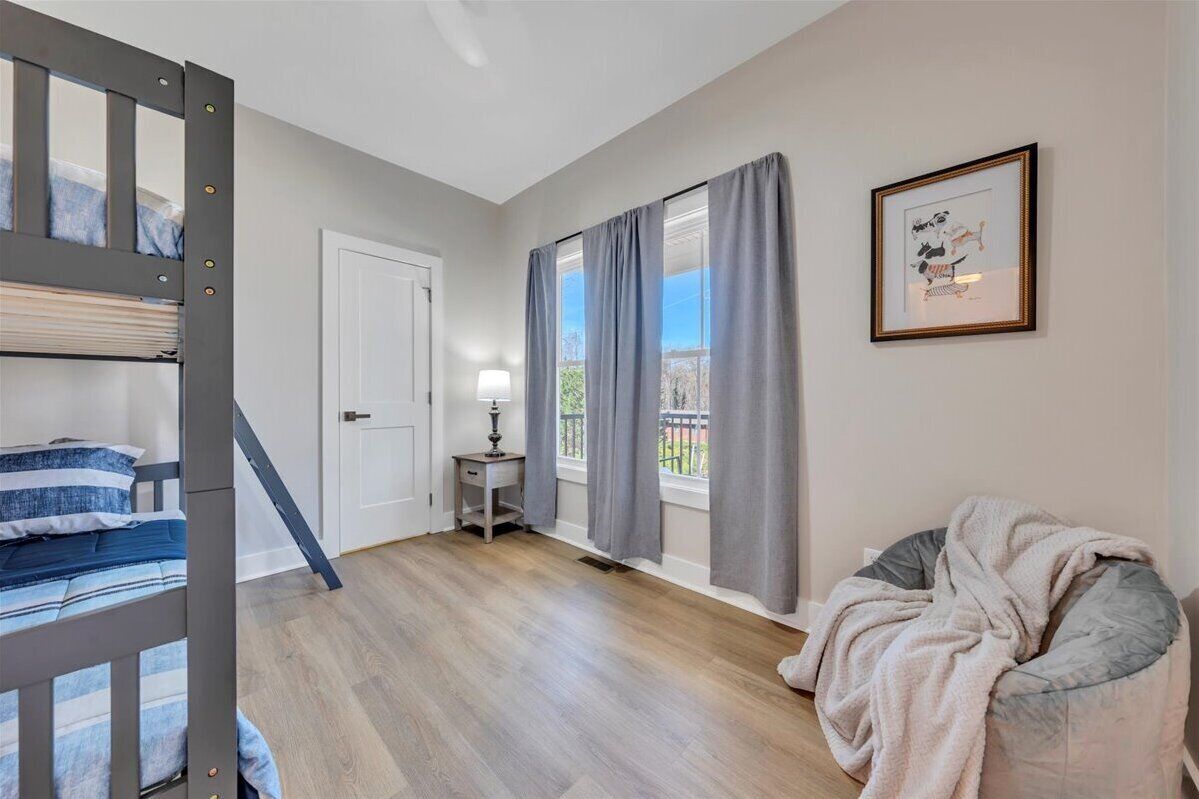 A room with a bunk bed, a bedside table with a lamp, a window with curtains, and a framed picture on the wall.