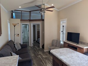 A spacious living area with a high ceiling, a ceiling fan, a large window with blinds, a dark-colored sofa with patterned cushions, a patterned armchair, a television on a wooden stand, and a kitchen area with a marble countertop. - The Most Beautiful Views in Turner Falls (Davis)