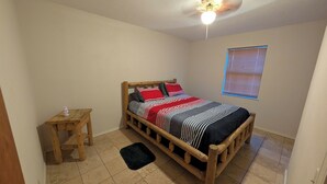 A bedroom with a wooden bed, a small wooden table, a black rug, a ceiling fan, and a window with blinds. - Mountain View 2 Bed Cabin (Davis)