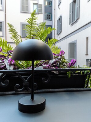 Une lampe de table noire sur un balcon doté d’une balustrade, entourée de plantes vertes et avec un bâtiment en arrière-plan.