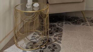 Un salon moderne avec un canapé beige, une table basse en verre aux pieds en fil d’or et un tapis à motifs.