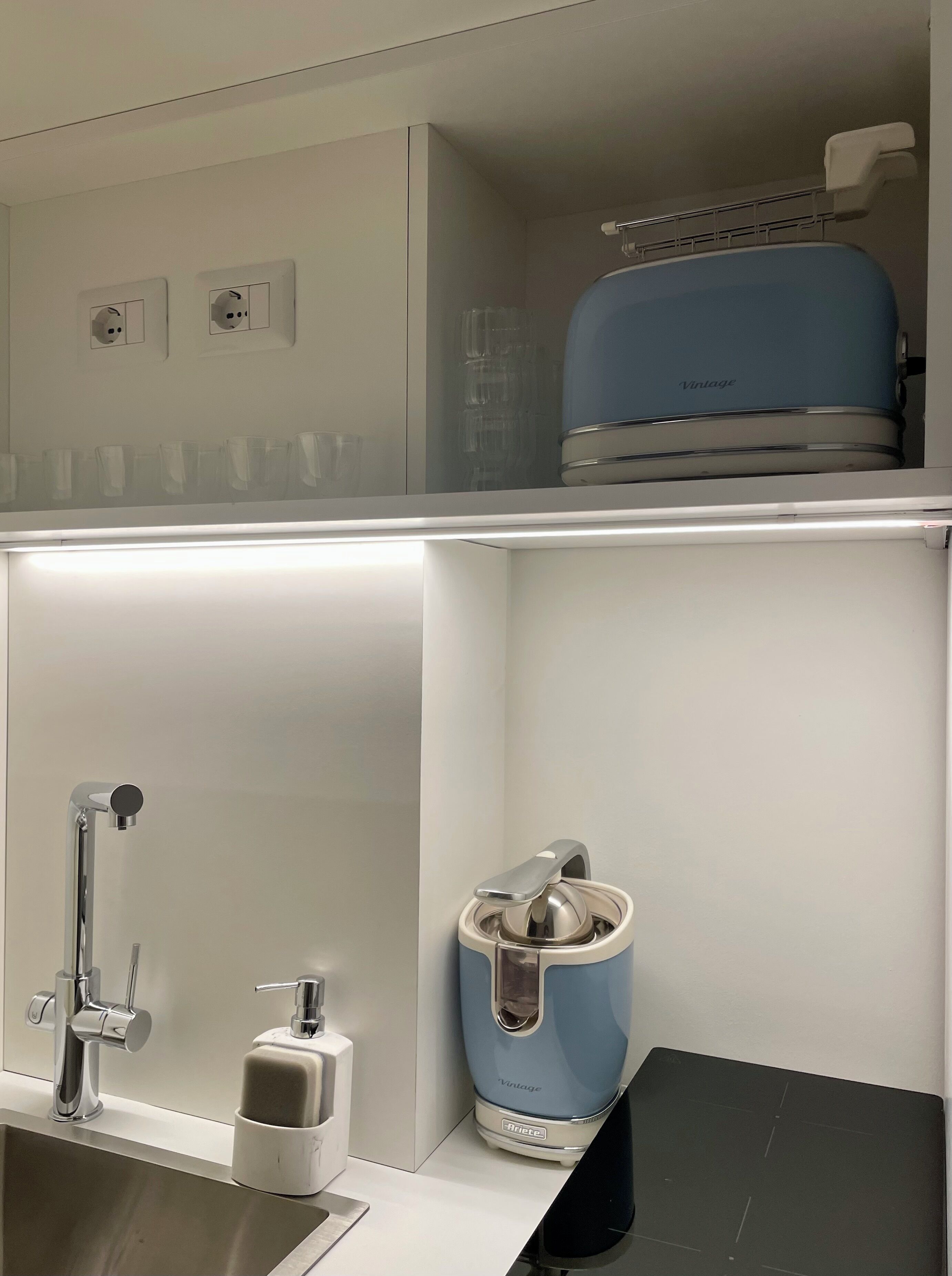 A modern bathroom with a stainless steel sink, a soap dispenser, a blue vintage-style electric toothbrush, and a glass shower enclosure.