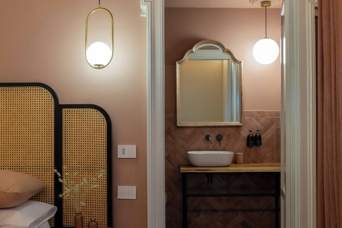 Une salle de bain moderne avec un grand miroir, un lavabo et des toilettes. On y trouve un lit avec une tête de lit et une petite table sur laquelle sont posés un vase et une bougie. Le sol est recouvert de carreaux décorés.